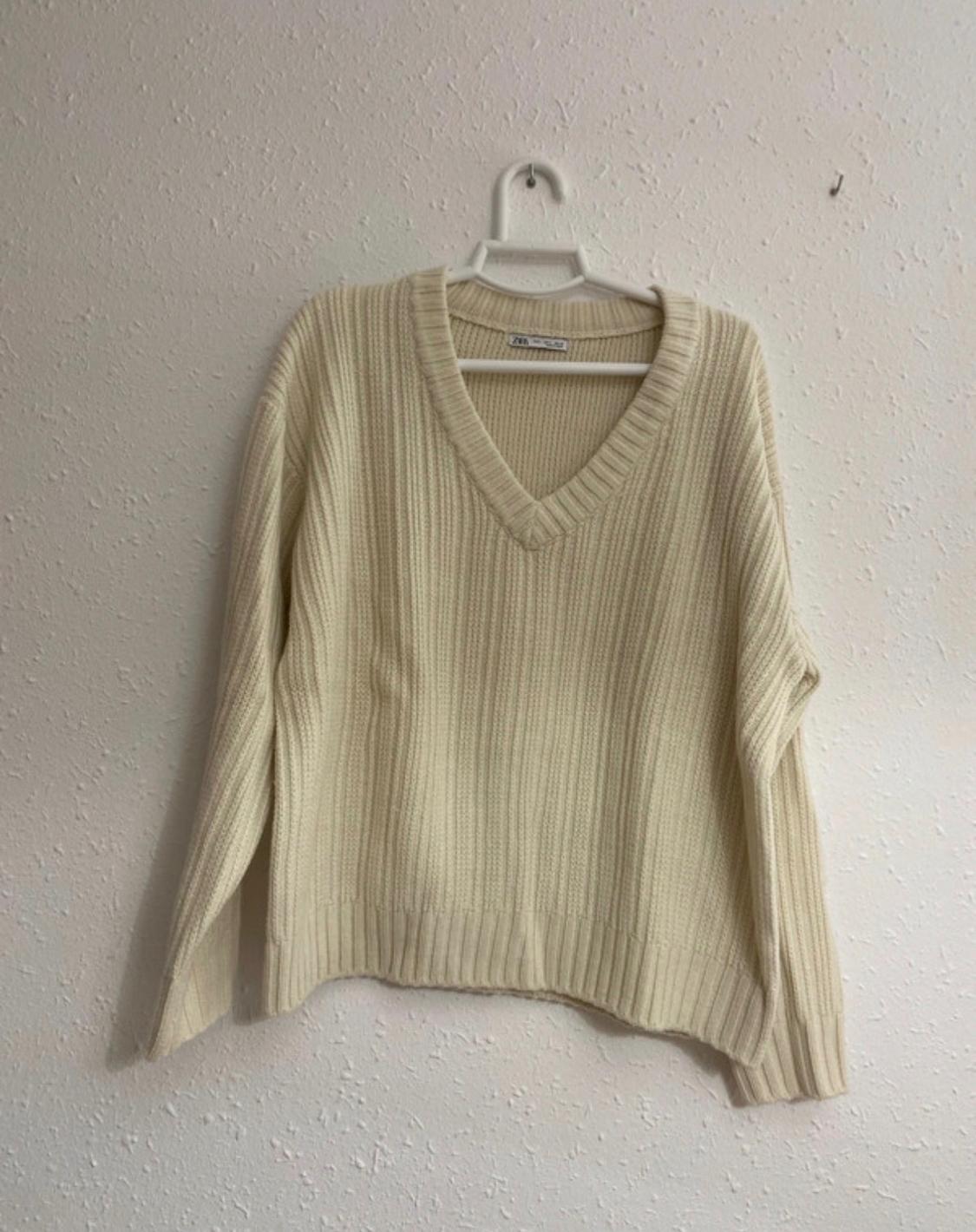 Cream ribbed-knit V-neck jumper, hanging on a white hanger against a textured wall. Label with ZARA text.