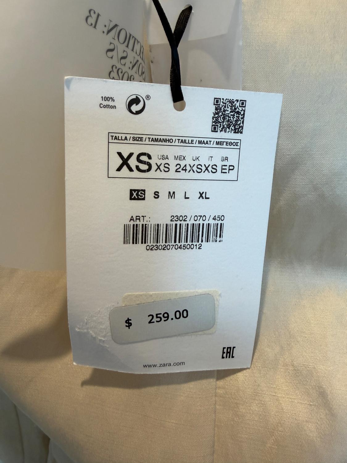Clothing tag with price $259.00, size XS, 100% cotton, barcode and QR. Beige fabric background. Text: www.zara.com.
