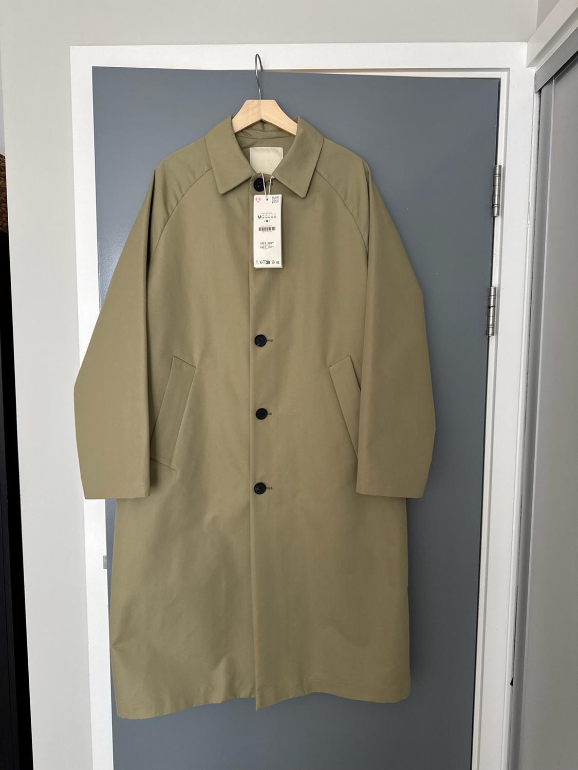 Long beige coat with a label, dark buttons and pockets, hanging on a wooden hanger over a grey door.