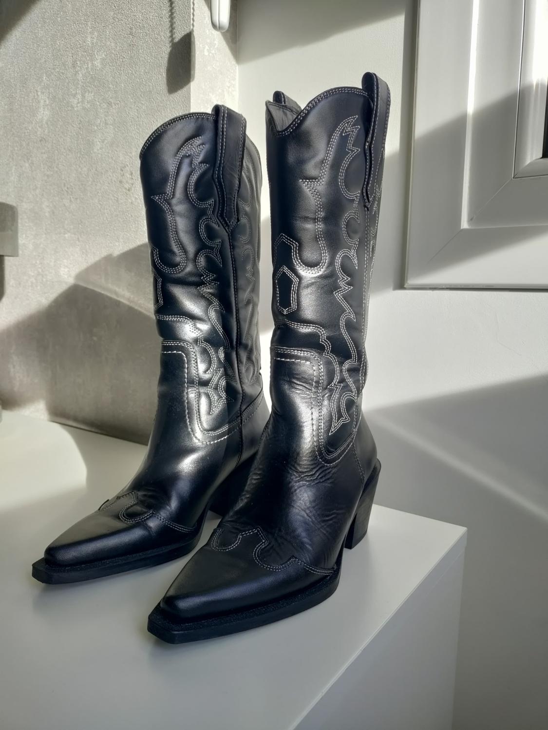 Black cowboy boots with decorative white stitching on a white surface, illuminated by natural light.