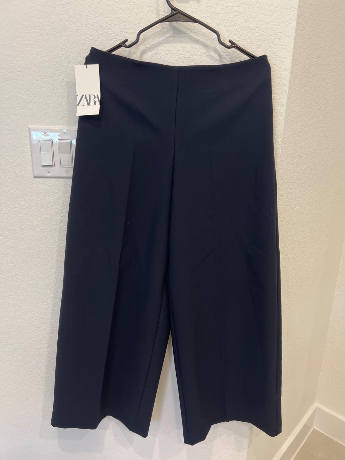Black pants hanging on a hanger with a ZARA tag, on a white wall next to a light switch.