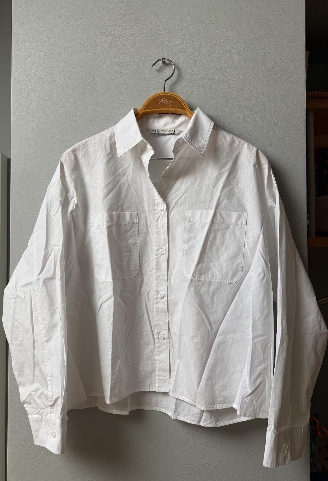 White long-sleeved shirt with front pockets, hanging on a wooden hanger against a grey wall.
