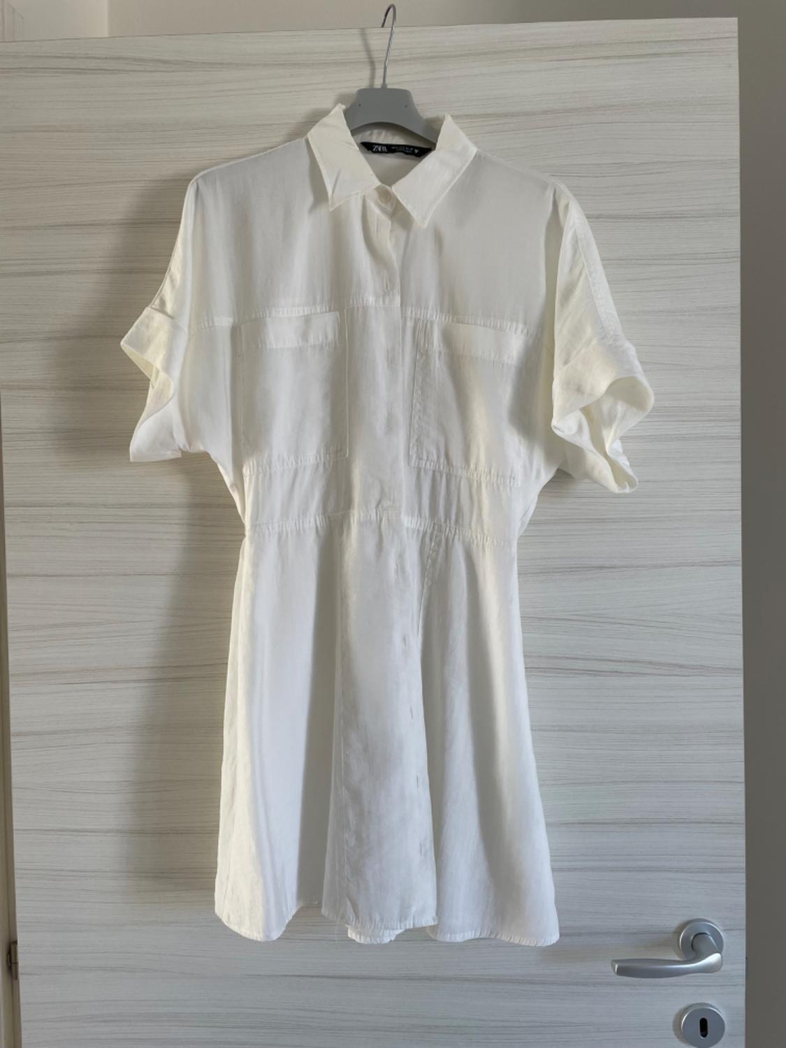 White short-sleeved shirt hanging on a hanger over a light wooden door. The shirt has a simple and elegant design.