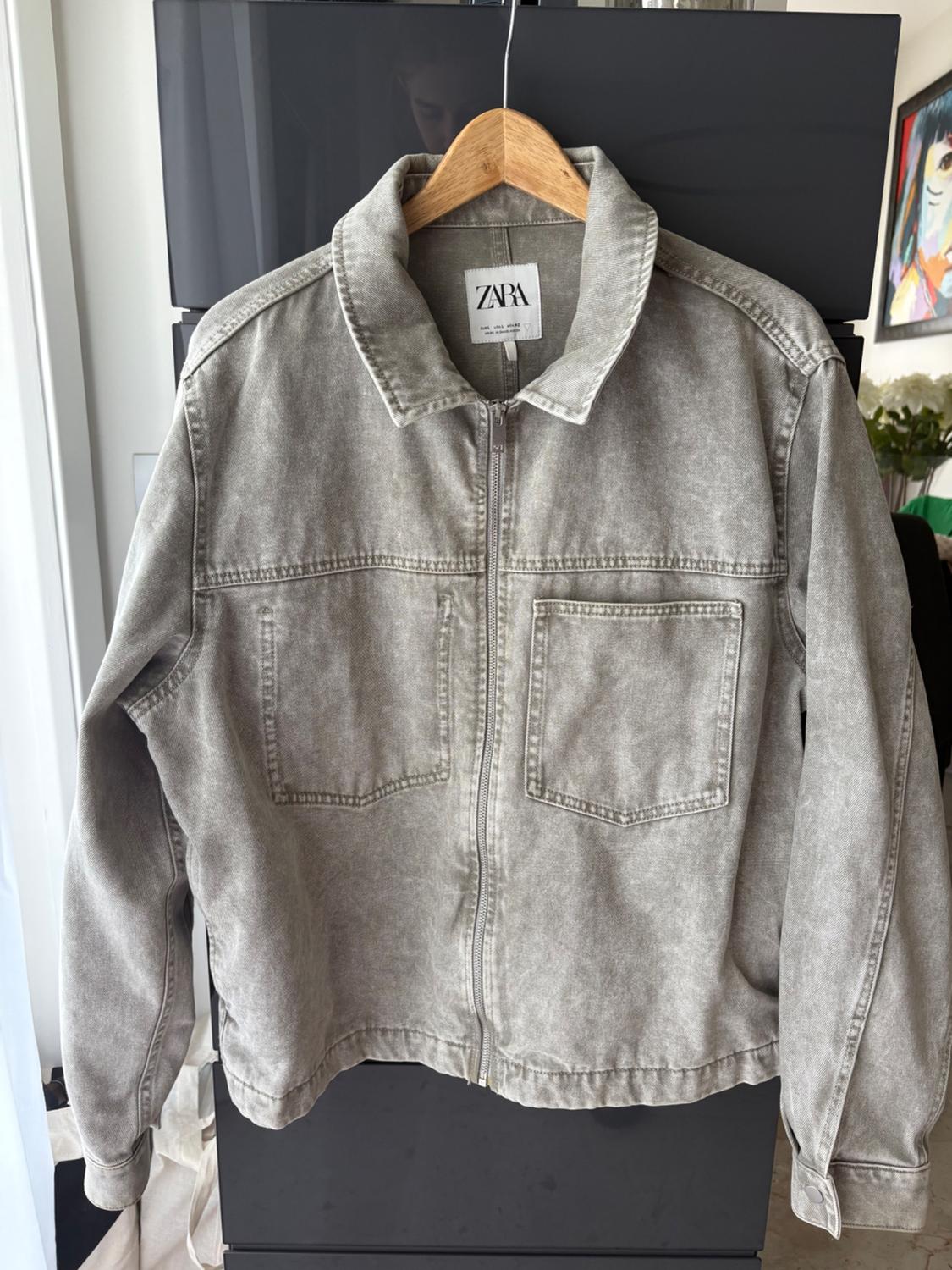 Grey denim jacket with a zip and two front pockets, hanging on a wooden hanger. Visible label: ZARA.