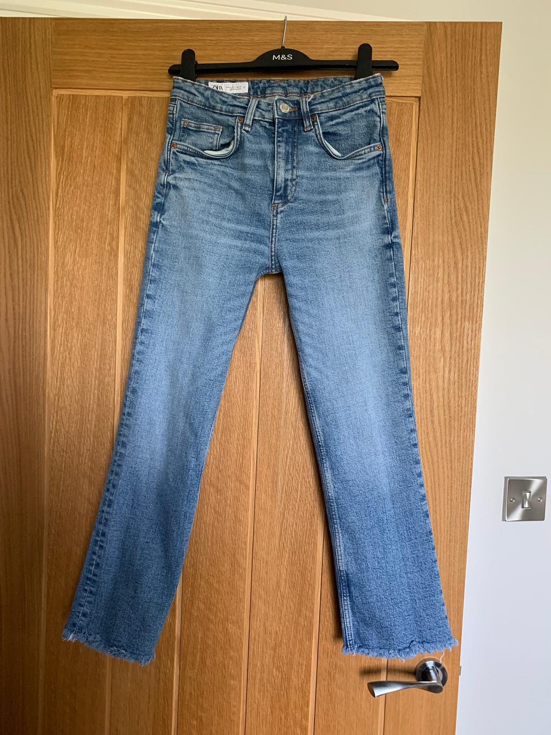 Blue jeans hanging on a black hanger over a wooden door. The hems have a frayed finish.