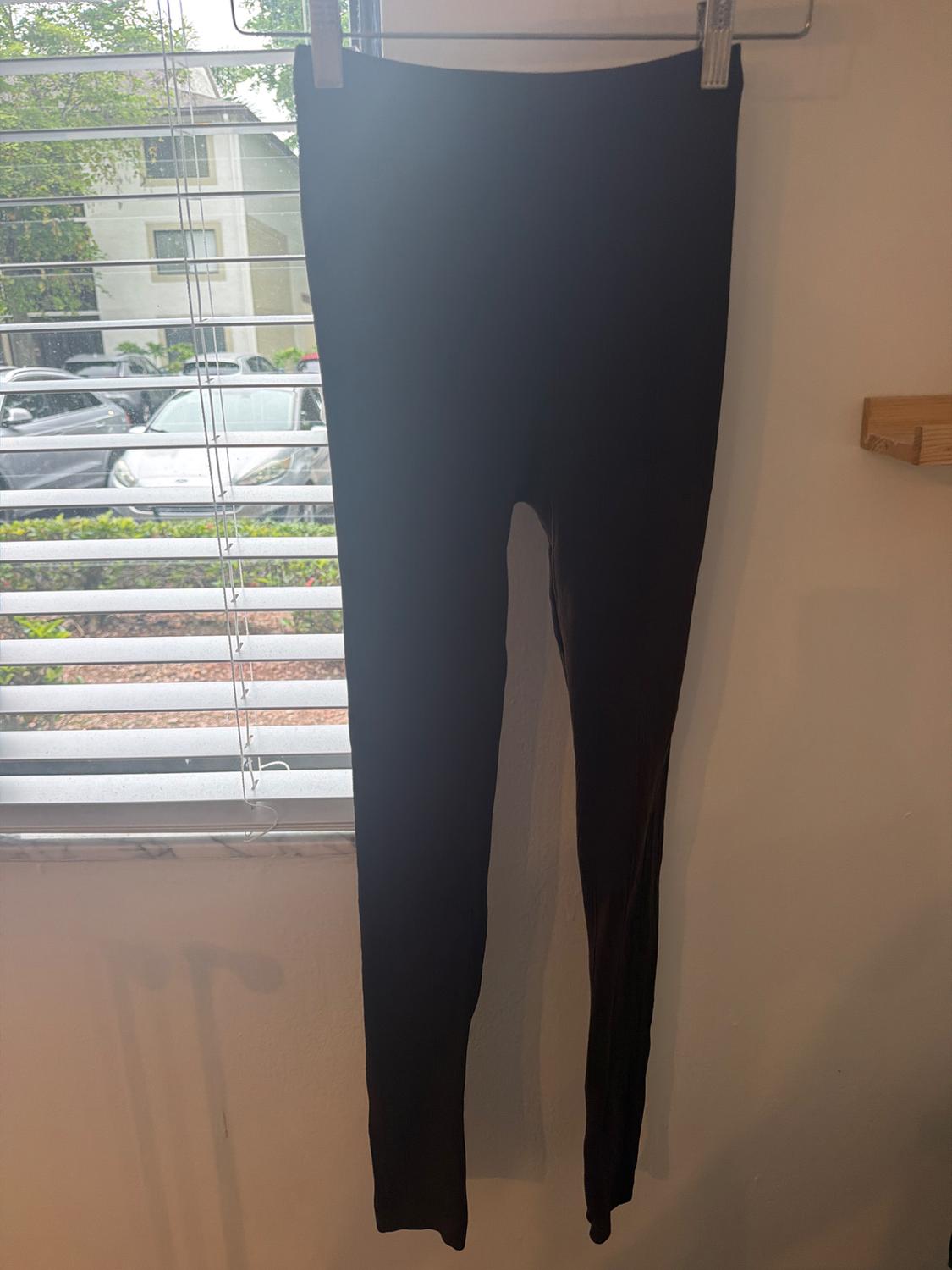 Black leggings hanging on a metal hanger on a white wall next to a window with blinds. Casual style.