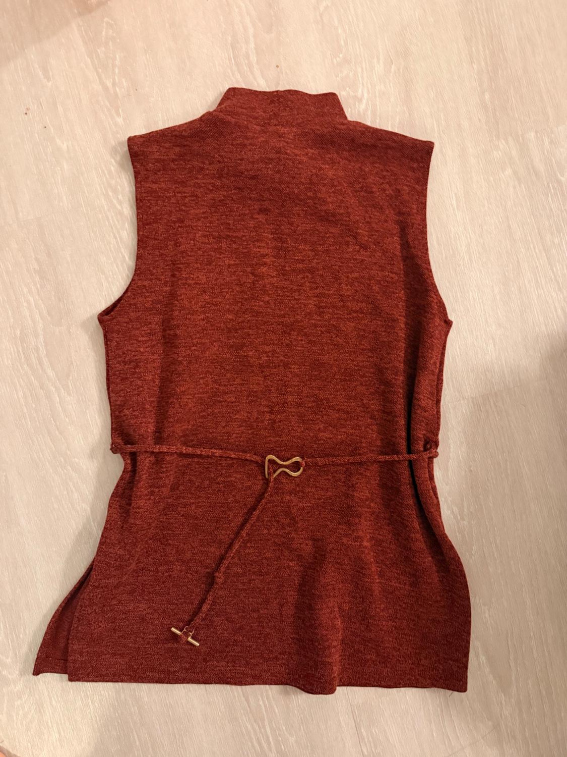 Sleeveless brick-colored high-neck top with a thin belt and gold detail, on a light wood background.