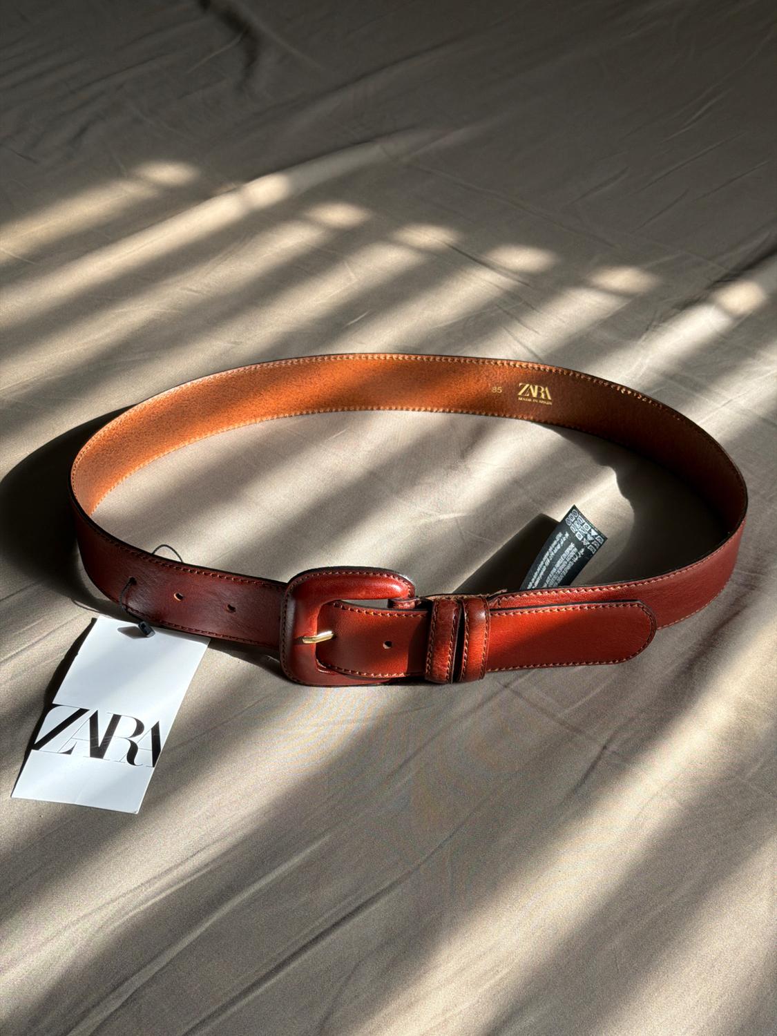 Brown leather belt with a gold buckle on beige fabric. Visible label with the text "ZARA". Light and shadows in the background.