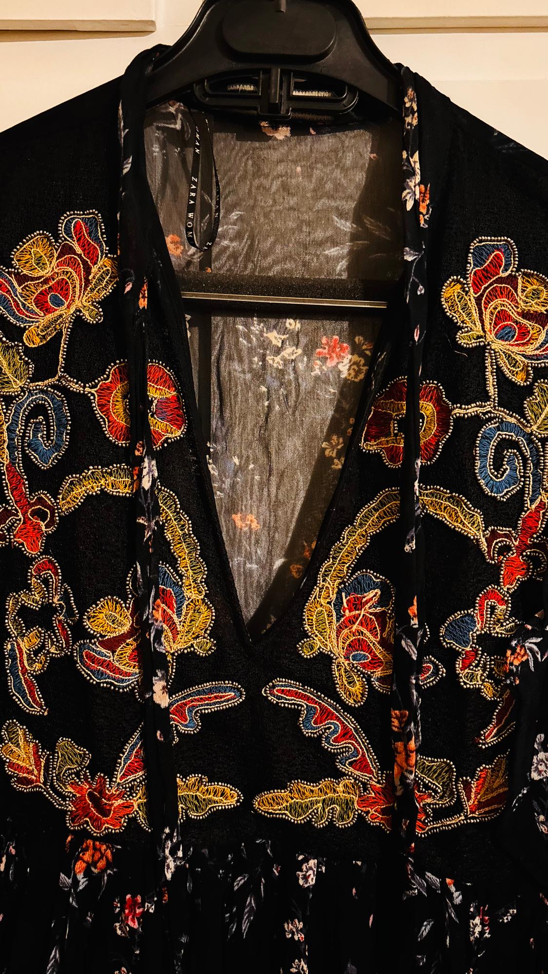 Black dress with floral embroidery in red, yellow, and blue, hanging on a black hanger. Visible label: "ZARA WOMAN".
