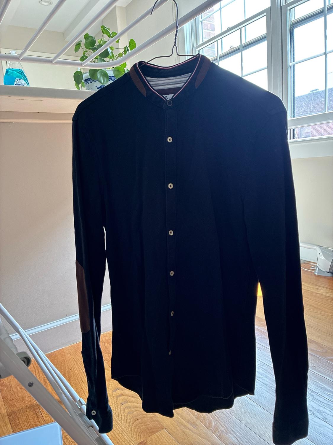Dark long-sleeve shirt with a Mandarin collar and brown elbow patches, hanging on an indoor clothesline next to a window.
