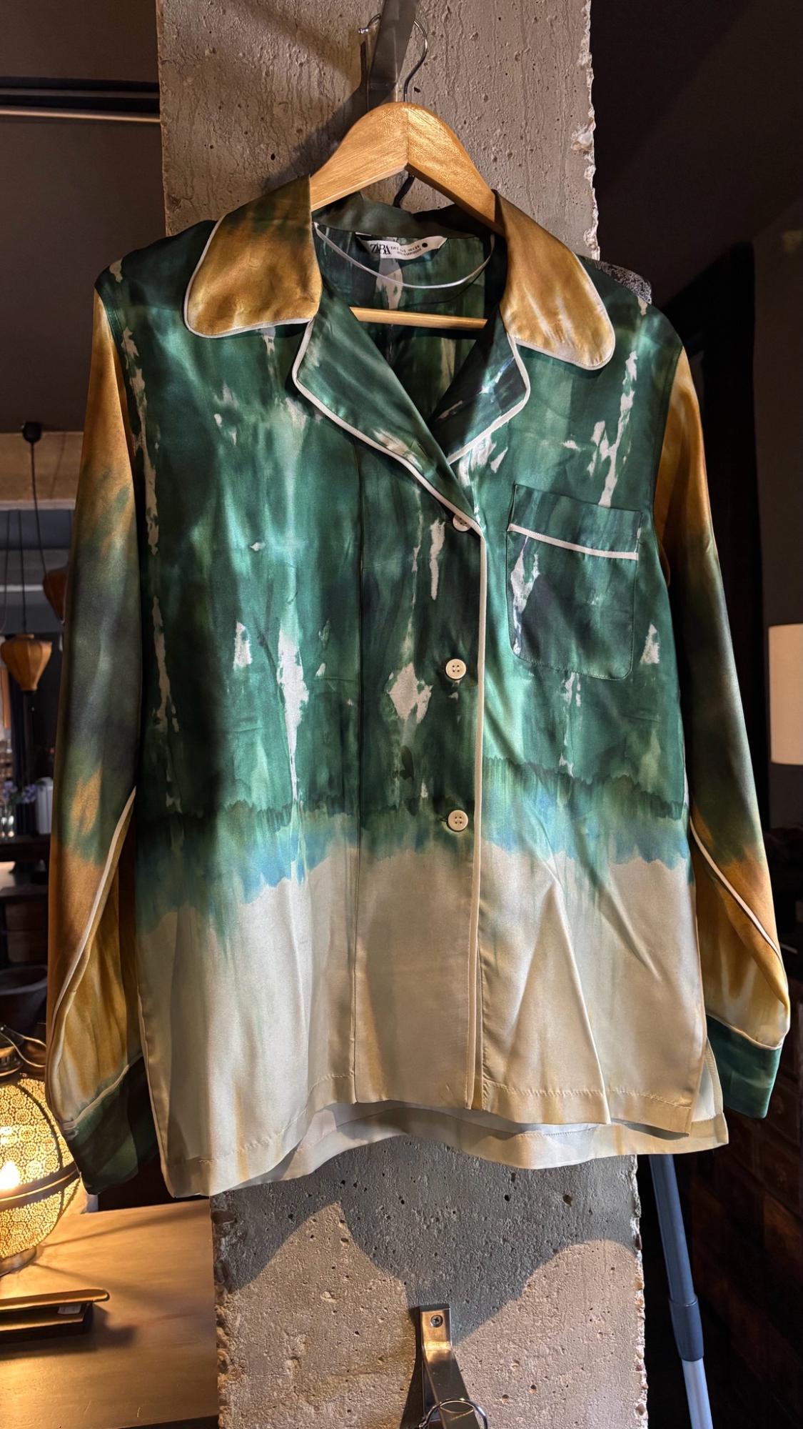 Green, white and ochre satin shirt with pocket, piping and ZARA label, hanging on a wooden hanger against a concrete pillar.