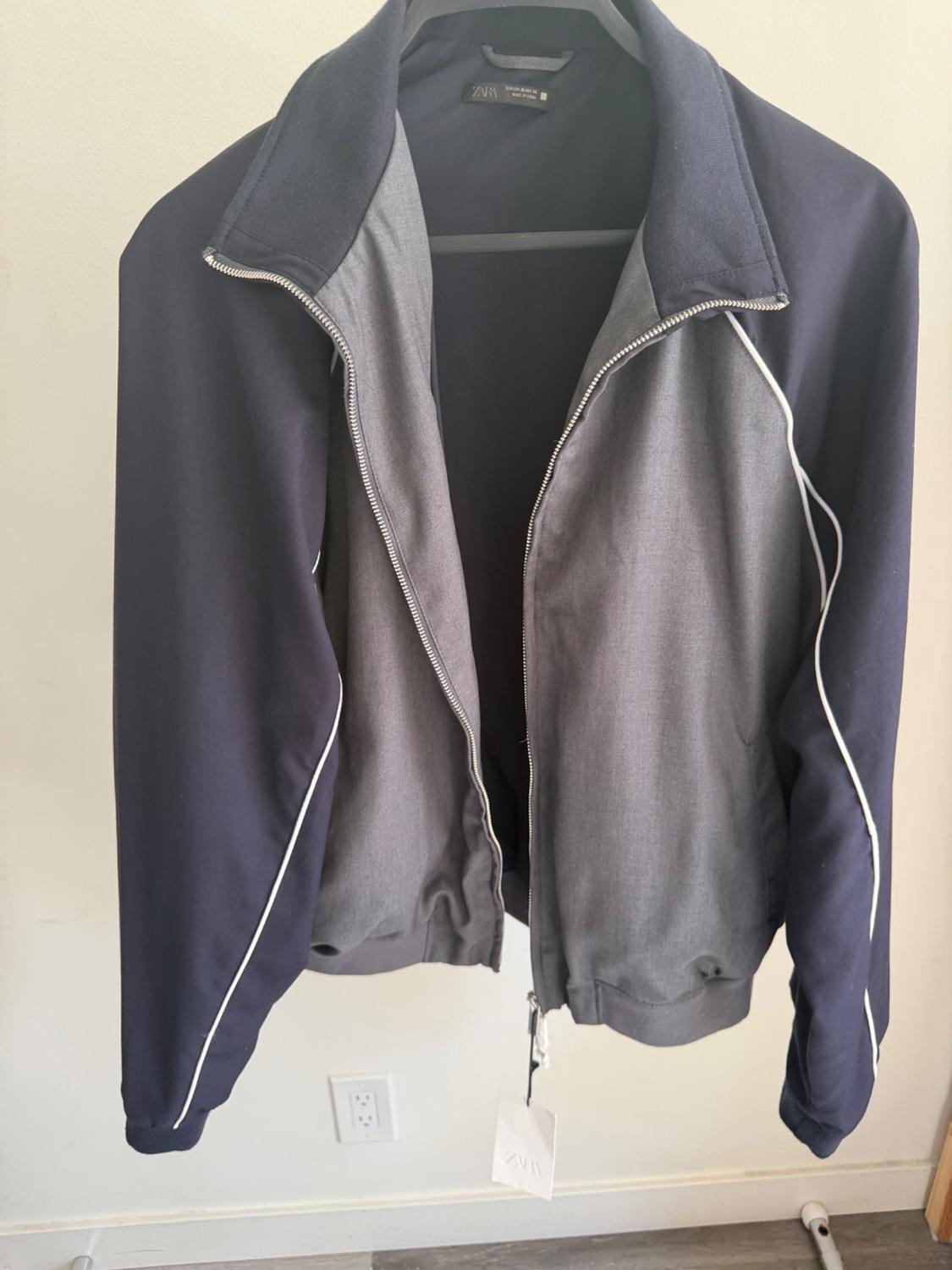 Jacket hanging on a hanger, with a grey body and dark blue sleeves with white trim. Visible label on the inside.