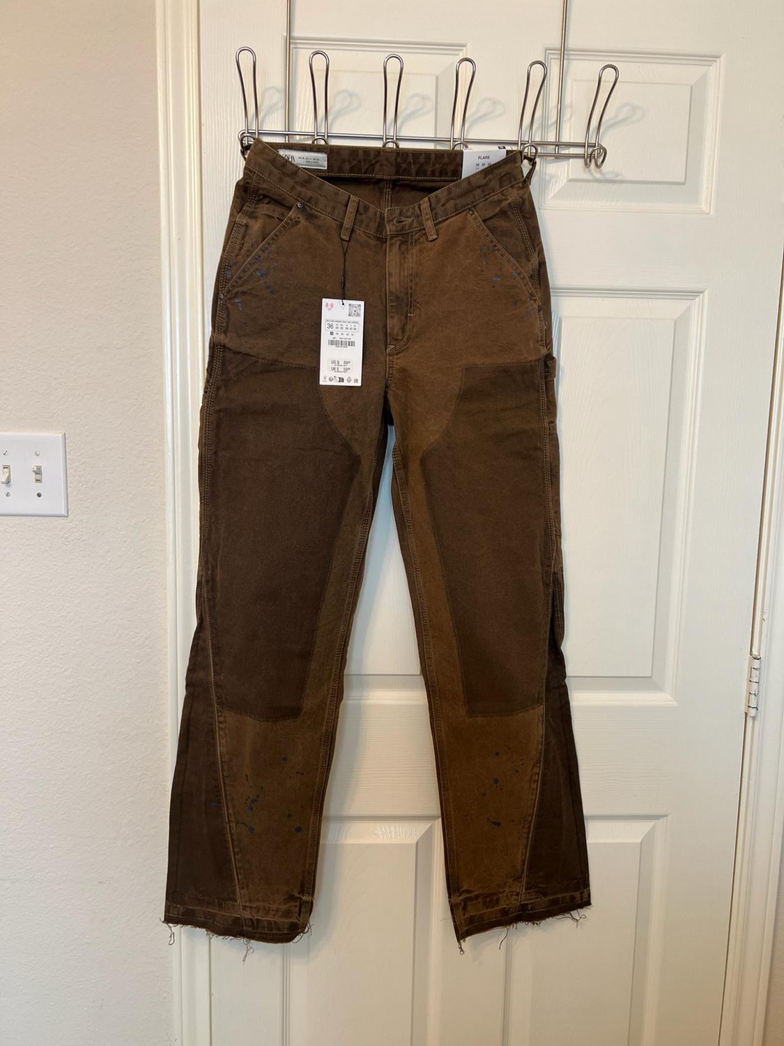 Brown trousers hanging on a white door. They feature dark panels, paint stains and a frayed hem. Visible text: 36, FLARE.
