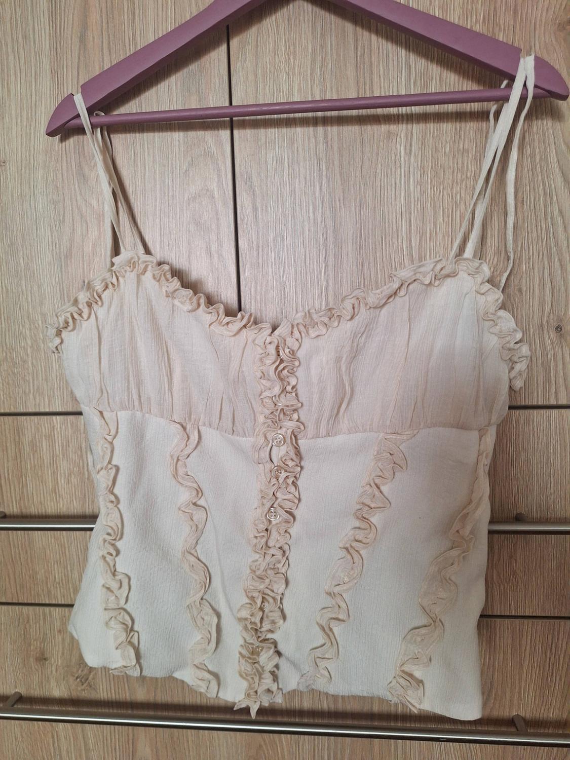 Beige camisole top with frills and front buttons, on a purple hanger against a wooden background. Romantic and relaxed style.