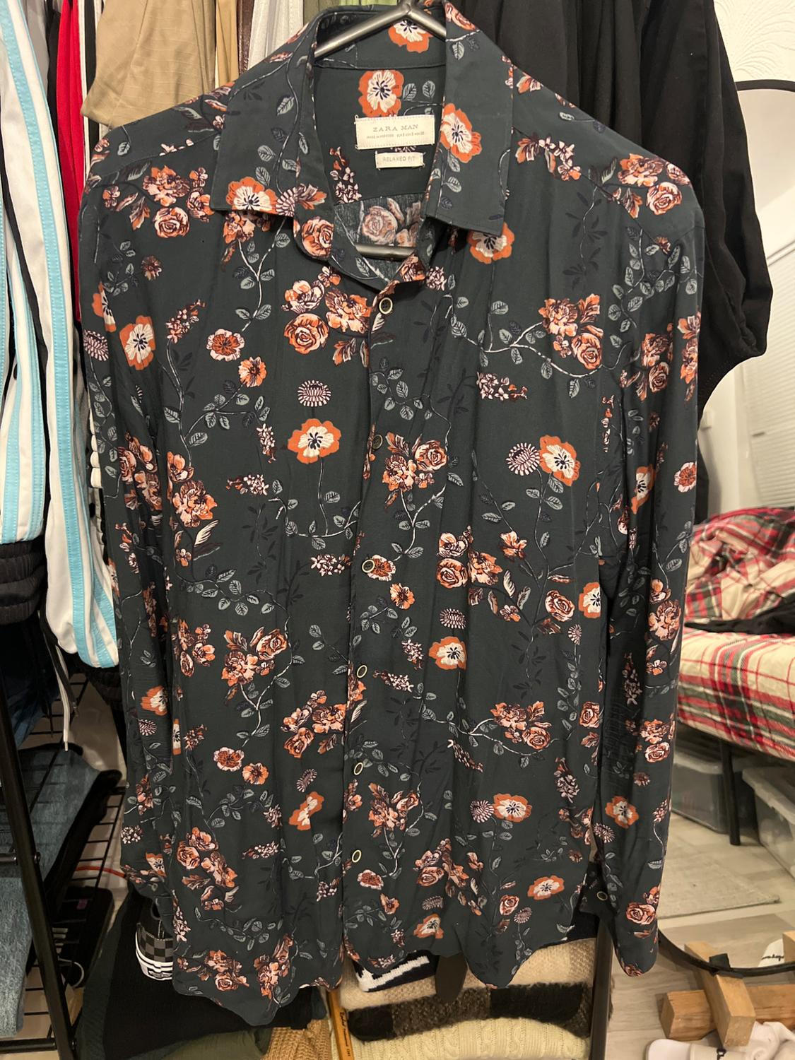 Men's floral print shirt in orange and white tones on a dark background, hanging in a wardrobe with assorted clothing.