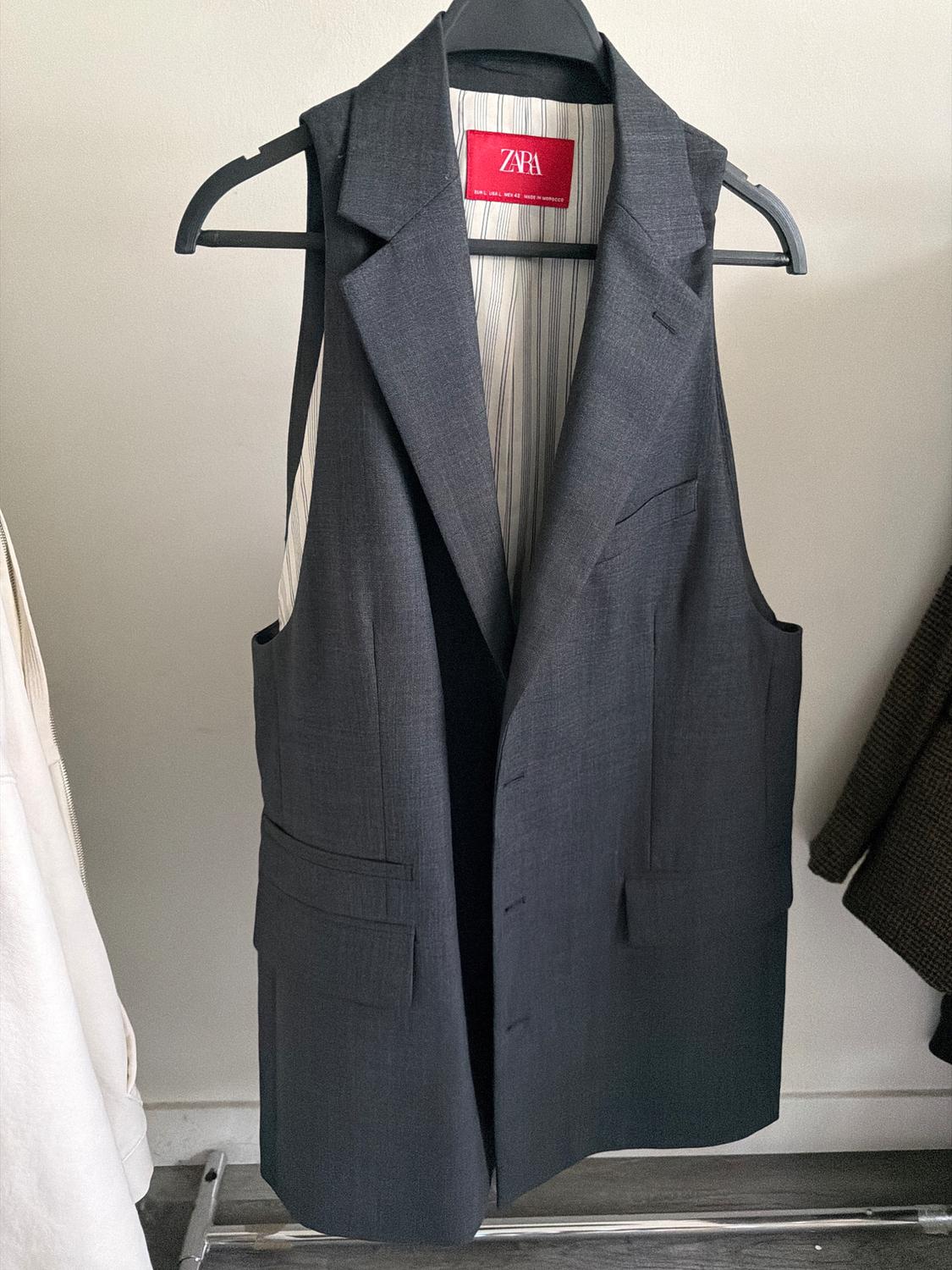 Grey sleeveless waistcoat hanging on a hanger, with a red Zara label visible inside. White wall background.