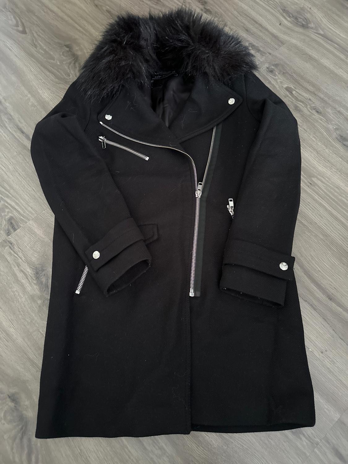 Black coat with asymmetric zip and faux fur collar on a grey wooden floor.