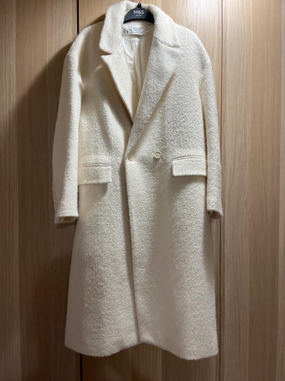 White wool coat hanging on a black hanger with an "M&S" tag. Light wood background.