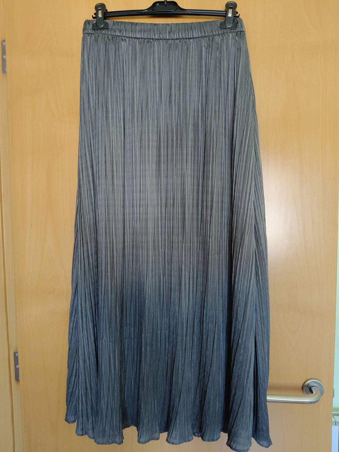 Long pleated skirt with a light to dark grey gradient, hanging on a black hanger over a wooden door with a silver handle.