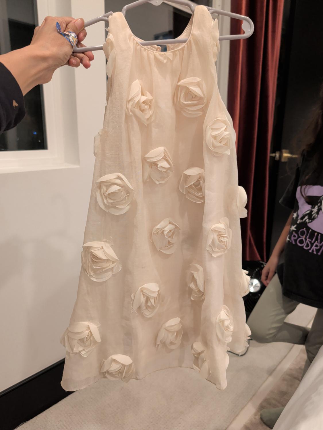 A white sleeveless dress with embossed flowers, held by a hand. The background shows red curtains and a white door.