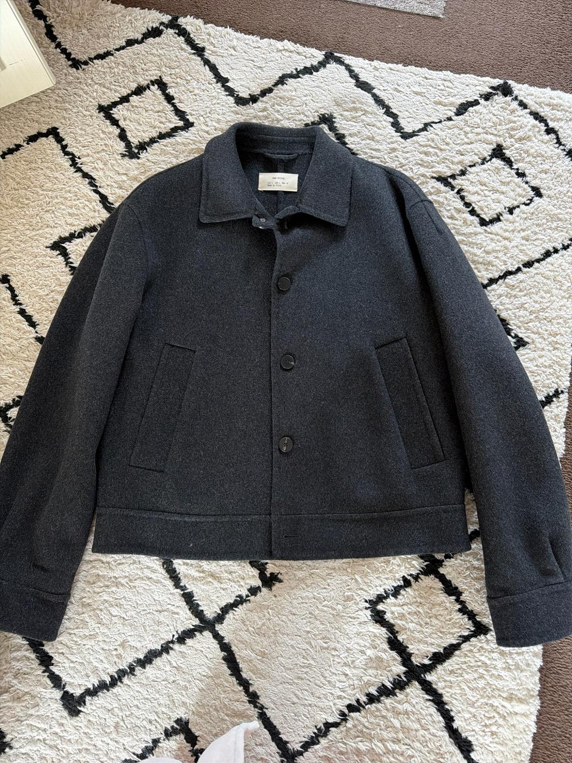 Dark grey button-up jacket with a collar and pockets, laid out on a soft-textured white rug with a black geometric pattern.