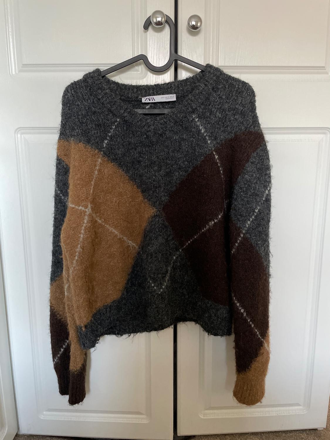 Knitted jumper with large grey, brown and camel argyle pattern, hanging on a hanger on a white door. Label with the text ZARA.