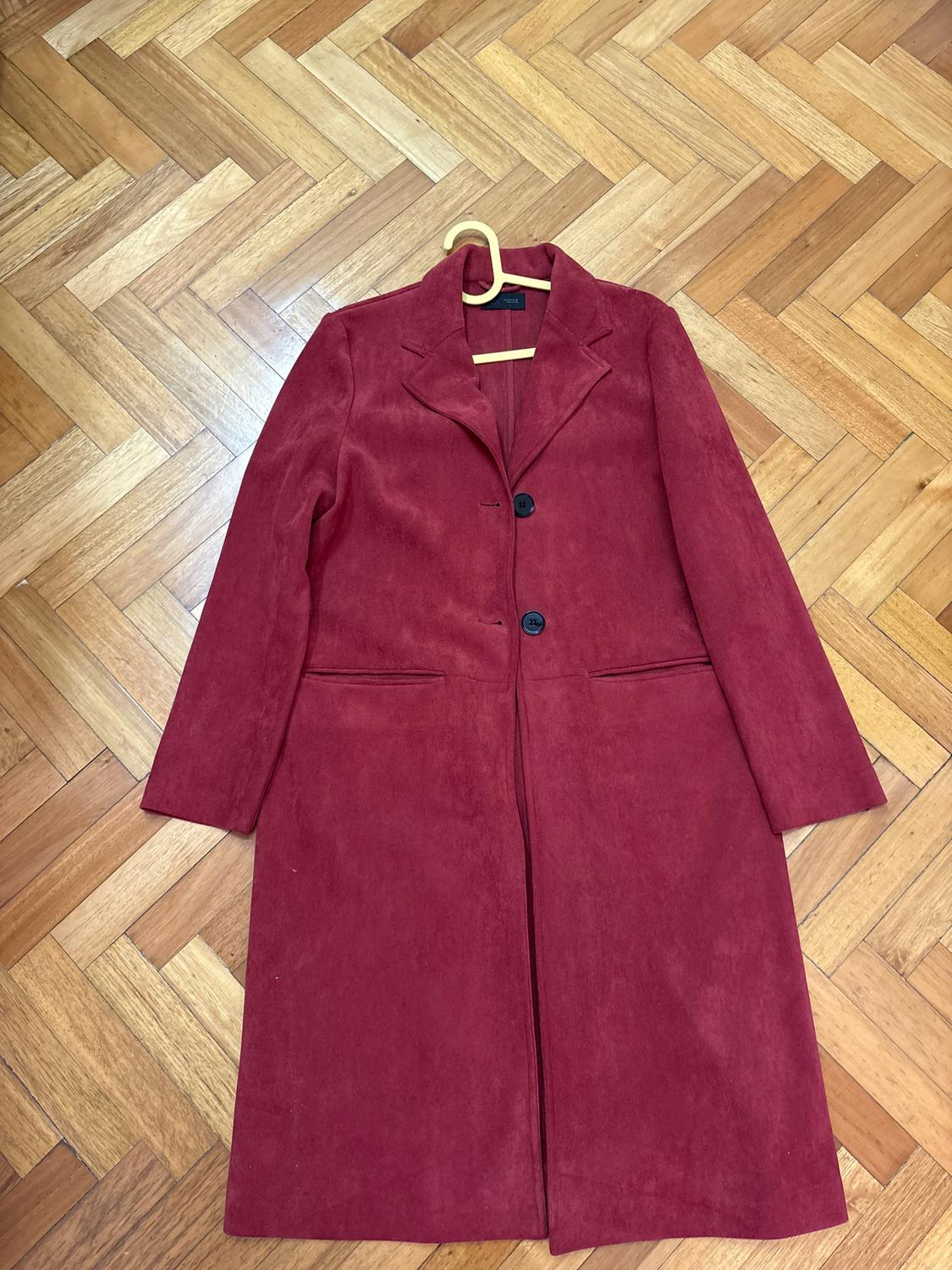 Long red coat with two black buttons, hanging on a hanger over a herringbone parquet floor.