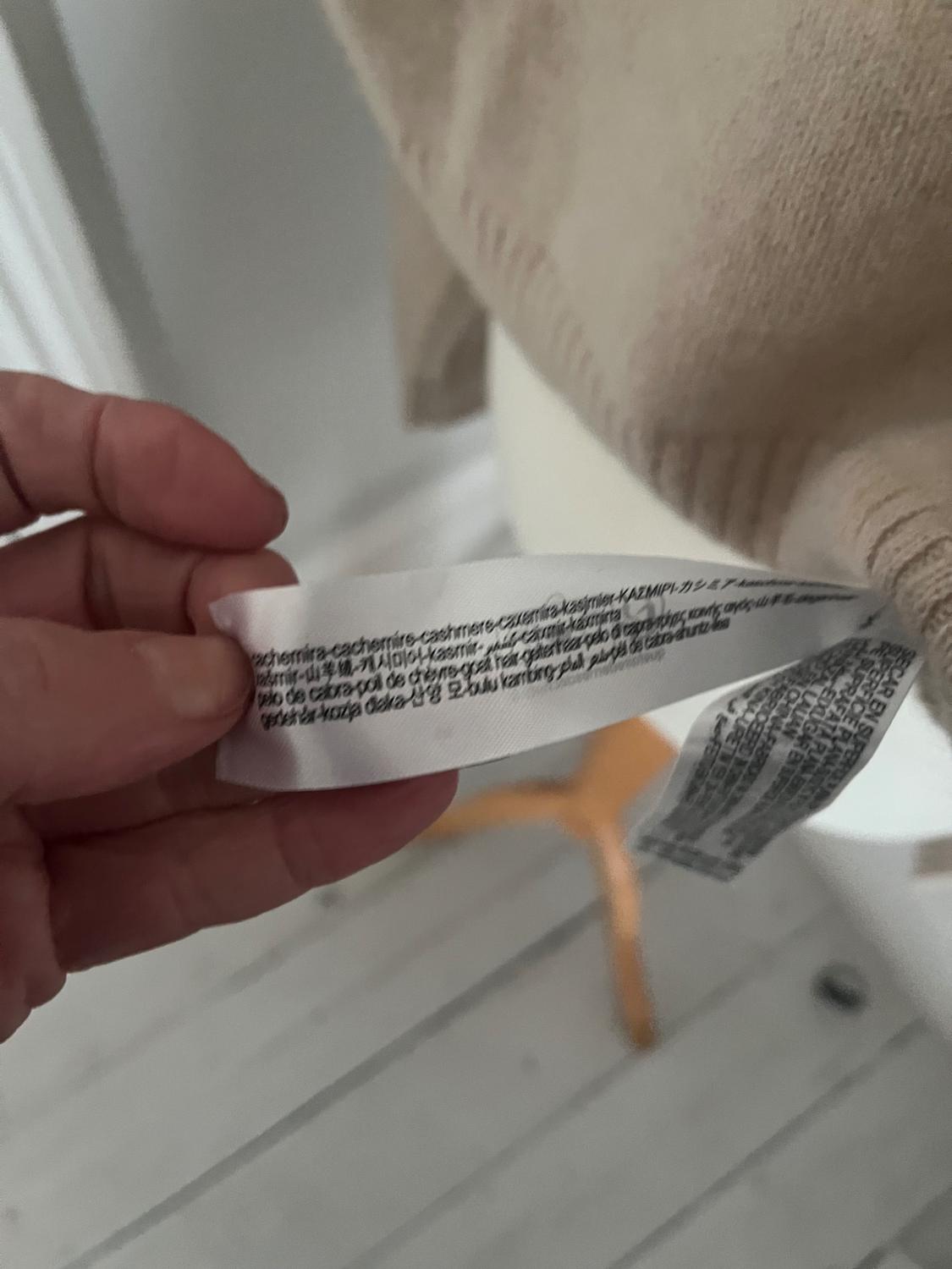 Hand holding a white fabric label with multi-language text. Blurred background shows part of a beige garment and wooden floor.