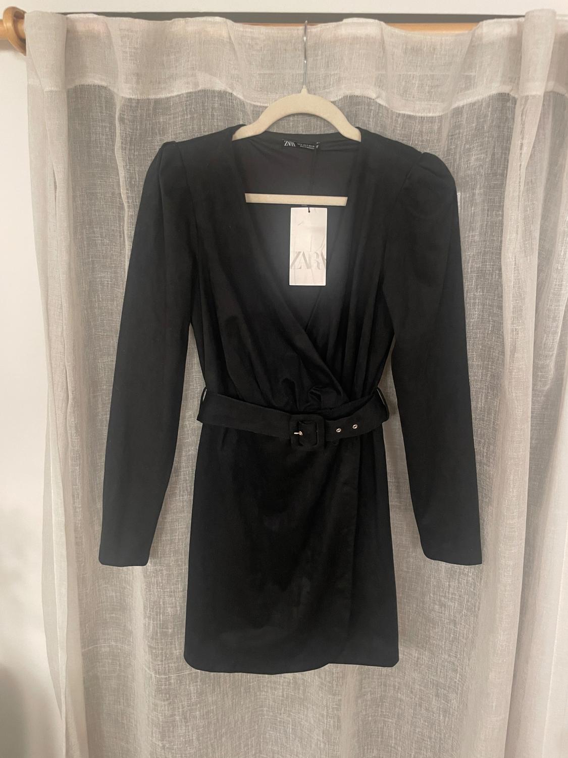 Long-sleeved black dress with belt, hanging on a hanger in front of a white curtain. Visible tag with the text "ZARA".