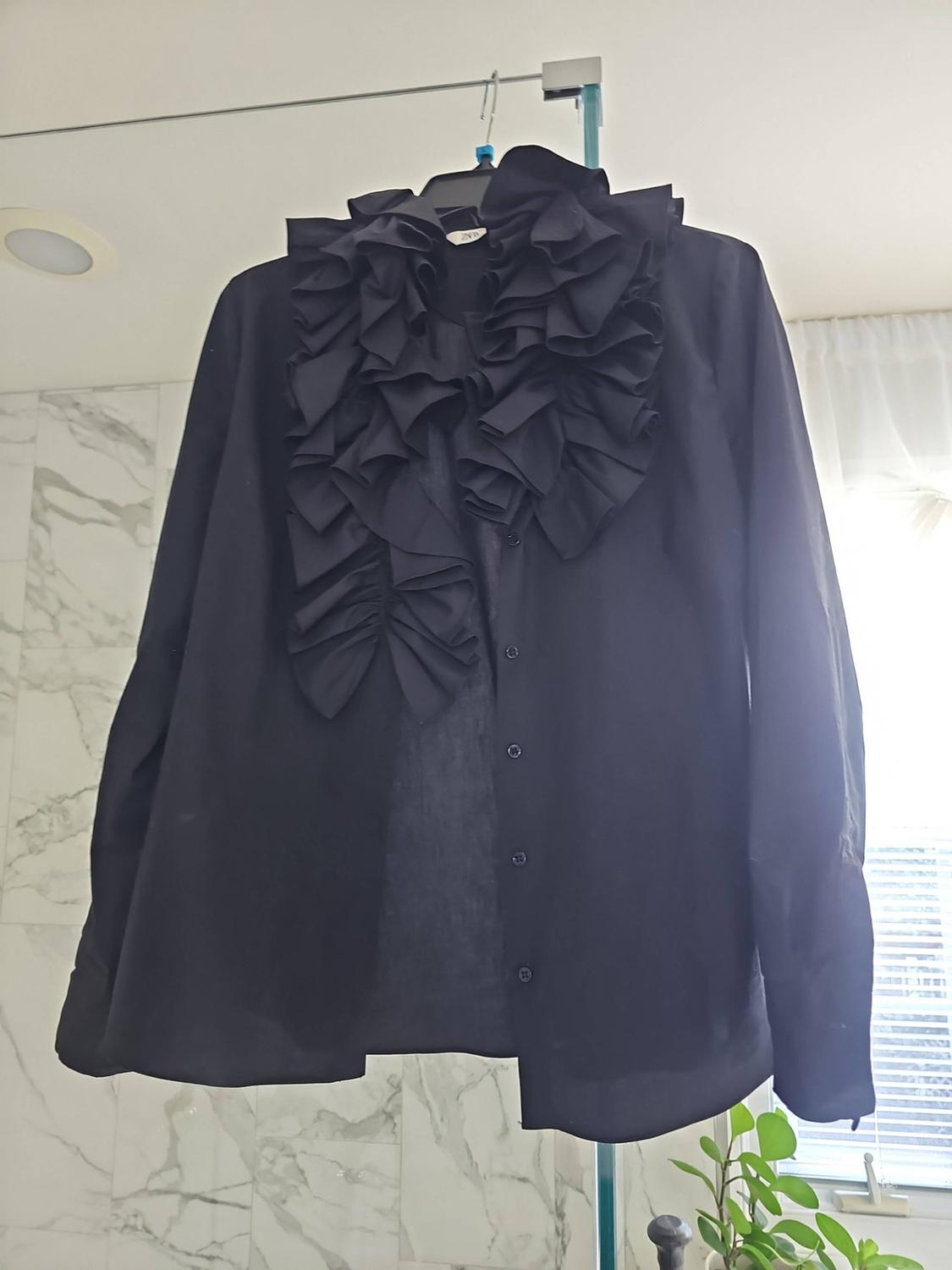 Long-sleeved black shirt with front ruffles, hanging on a hanger in a bright bathroom with marble-effect tiles and a plant.