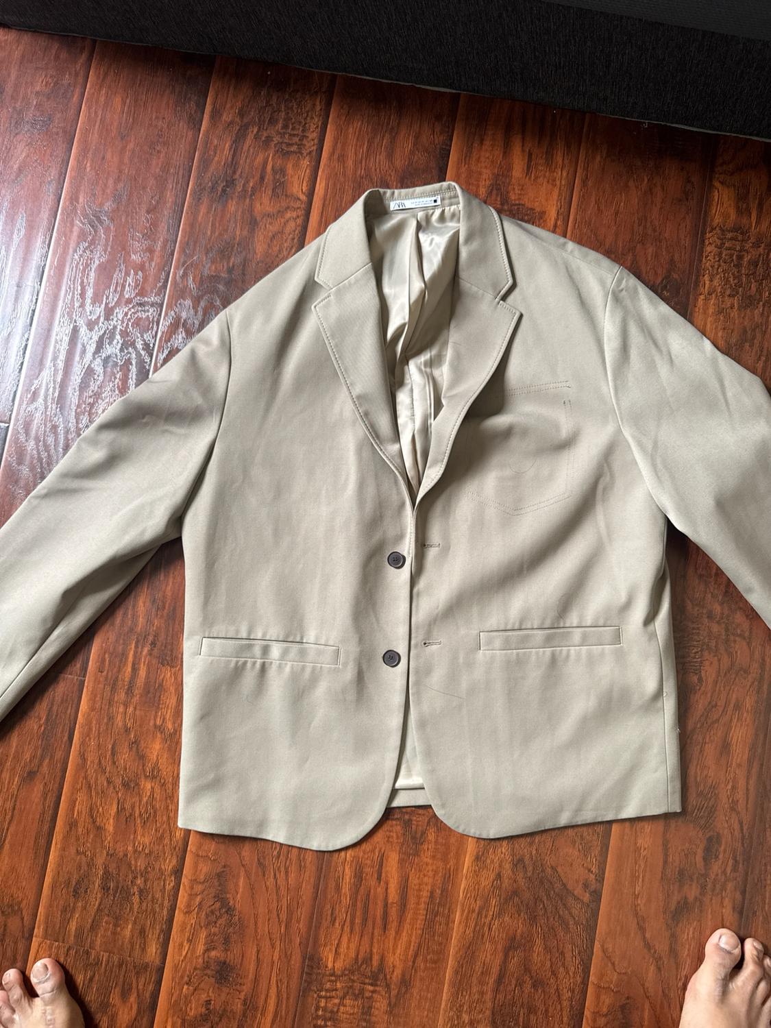 Beige jacket with two buttons and pockets on a wooden floor. ZARA label visible. Bare feet protrude at the bottom.