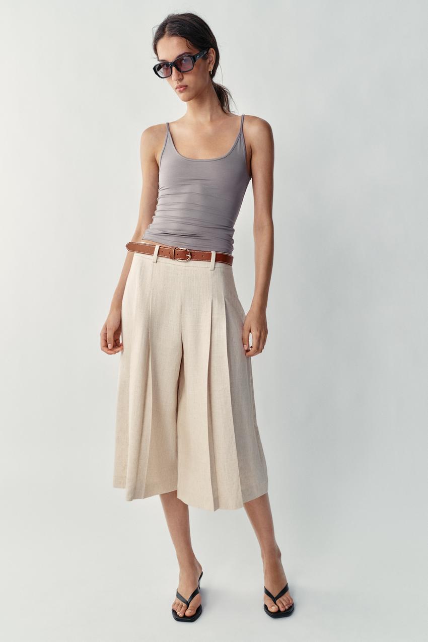 Conjunto de top gris de tirantes, bermuda larga beige claro con pinzas delanteras y cinturón marrón, gafas de sol y sandalias negras.