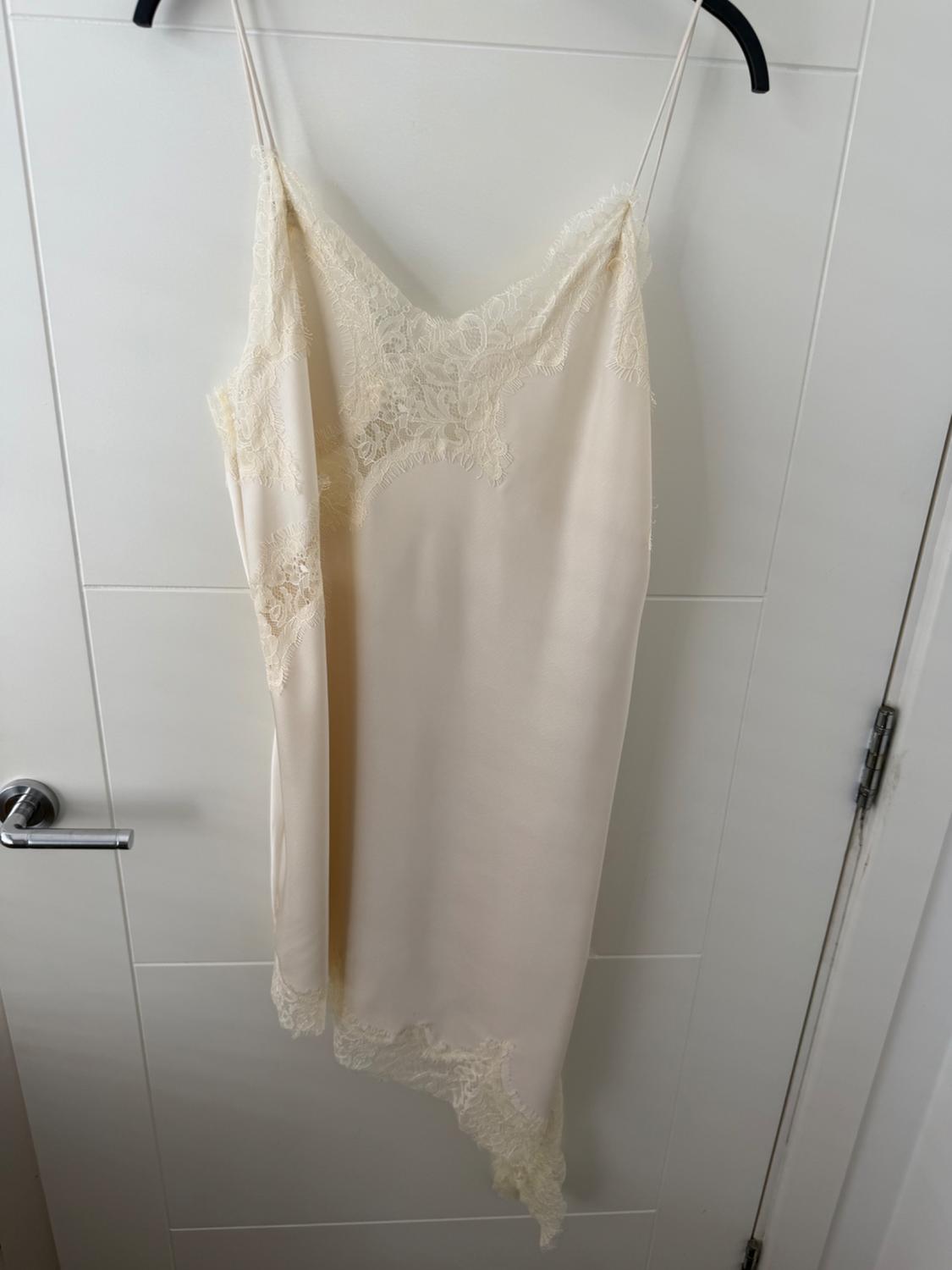 Cream satin slip dress with thin straps, lace, and an asymmetrical hem, hanging on a black hanger over a white door.