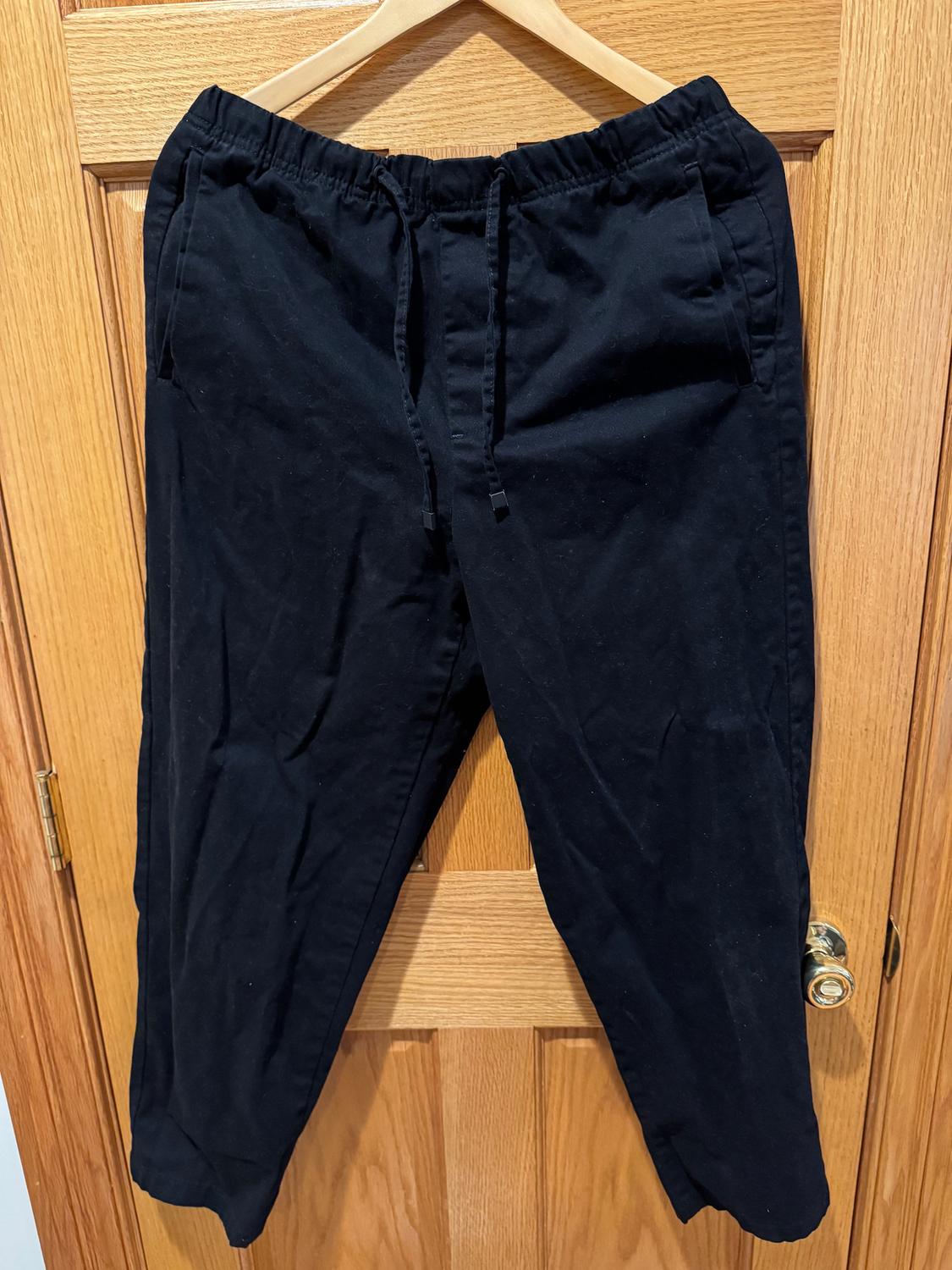 Black trousers hanging on a wooden hanger over a light wooden door. Elasticated waist with adjustable drawstring.