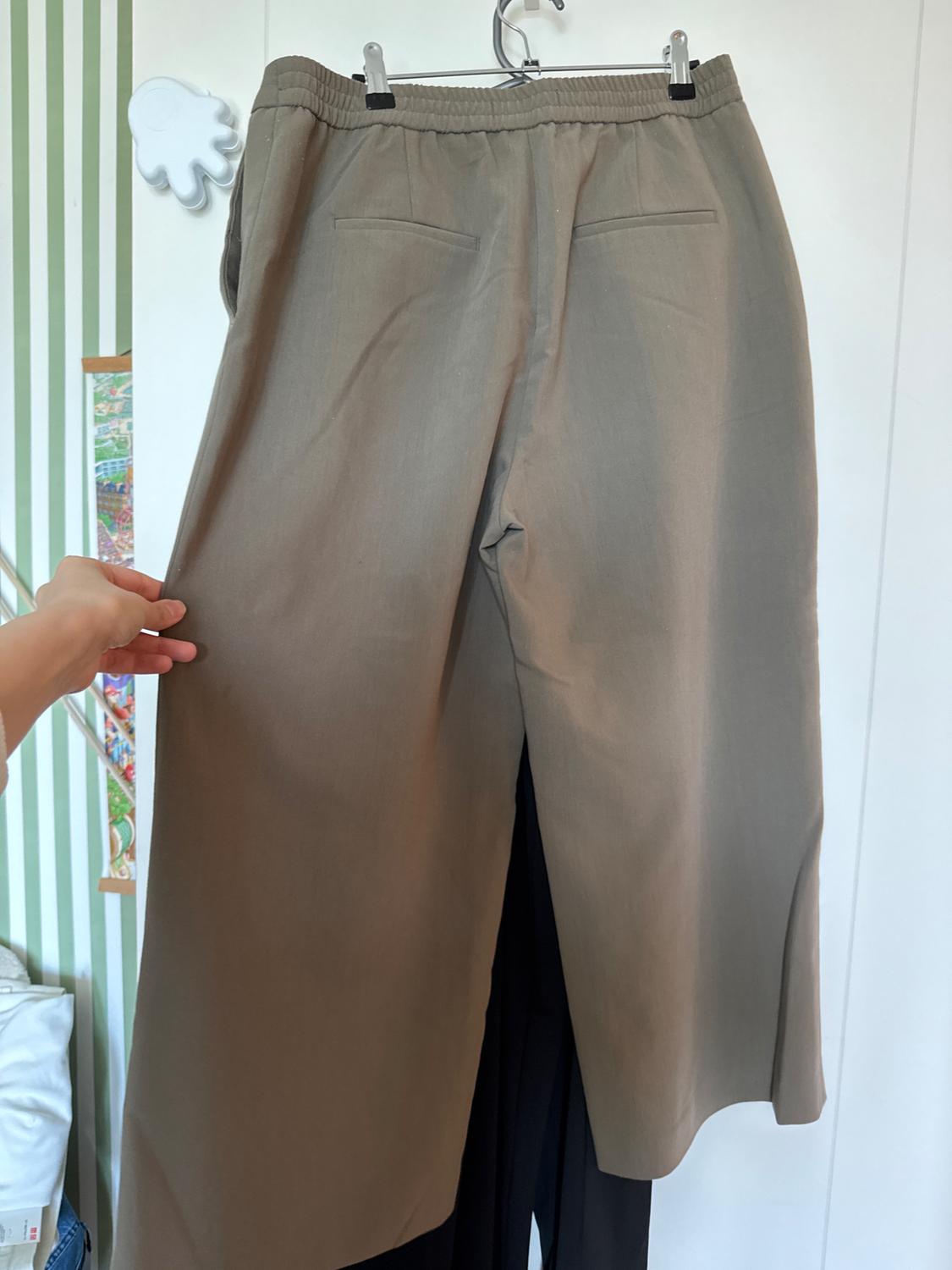 A pair of beige trousers hanging on a hanger, held by a hand. The background features green and white stripes along with a wall decoration.