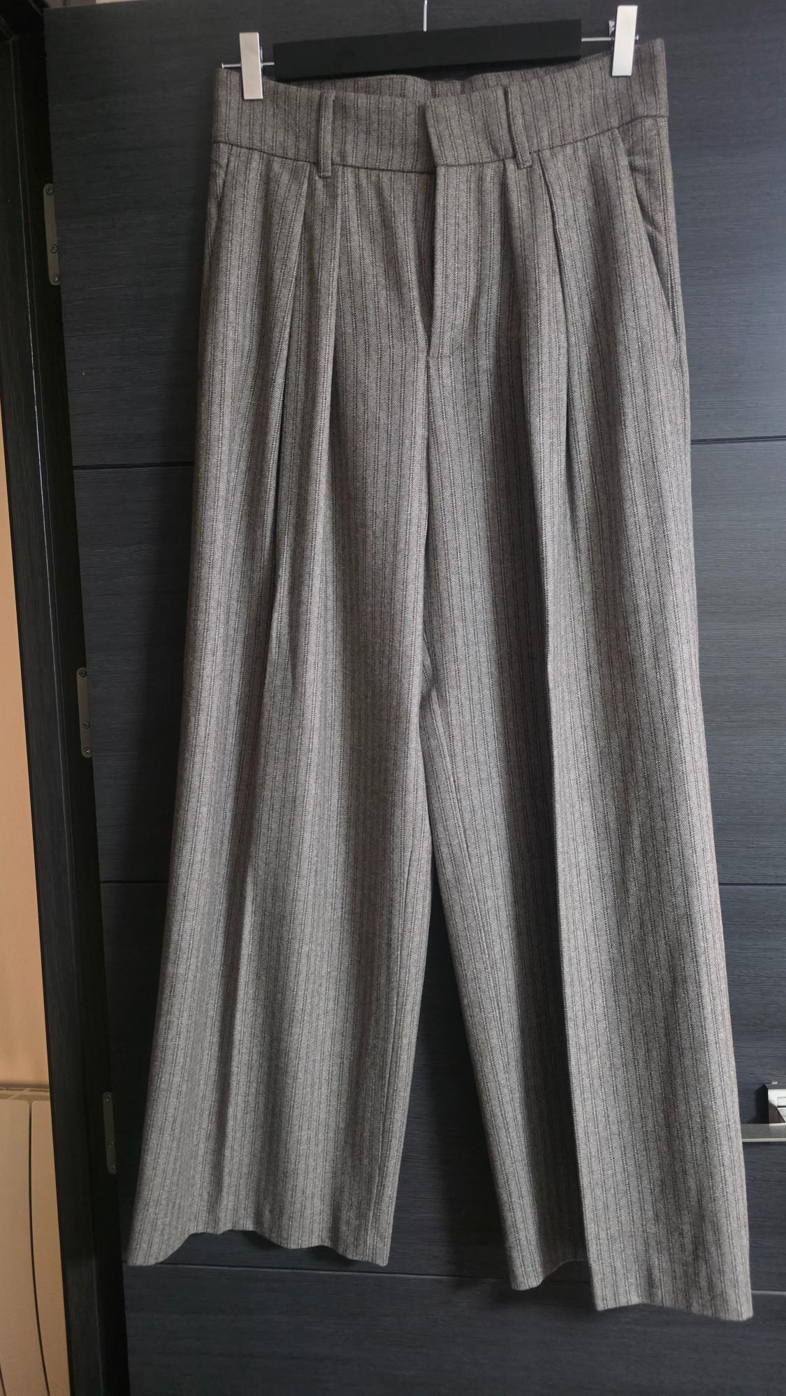 Grey wide-leg pinstripe trousers with pleats, hanging on a black hanger in front of a dark door.