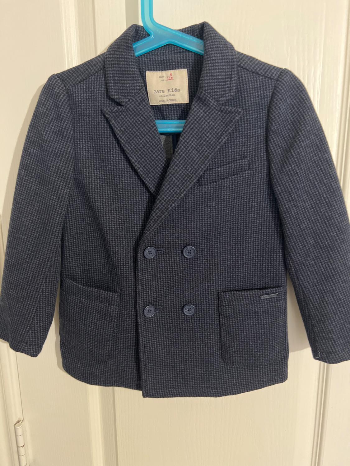 Dark blue children's jacket with a small plaid print, double-breasted, and pockets, hanging on a blue hanger. Label: Zara Kids.