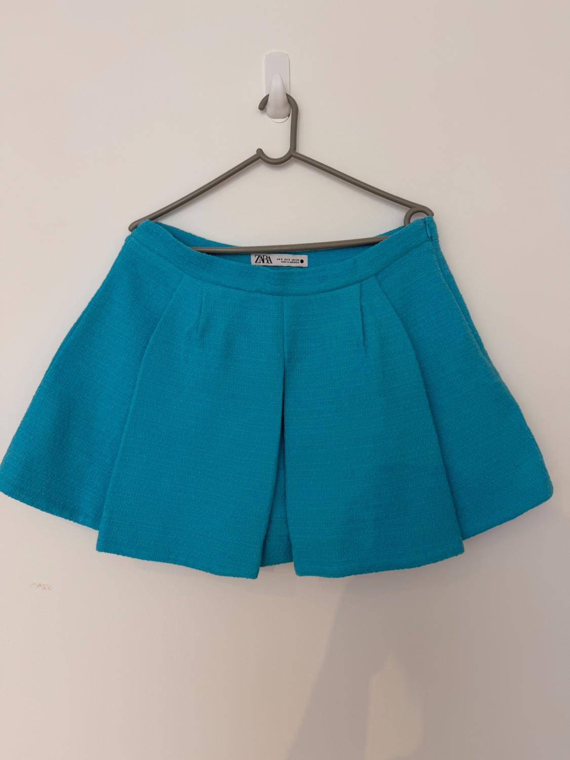 Turquoise blue pleated mini skirt, hanging on a grey hanger against a white wall. Interior label visible with the text ZARA.