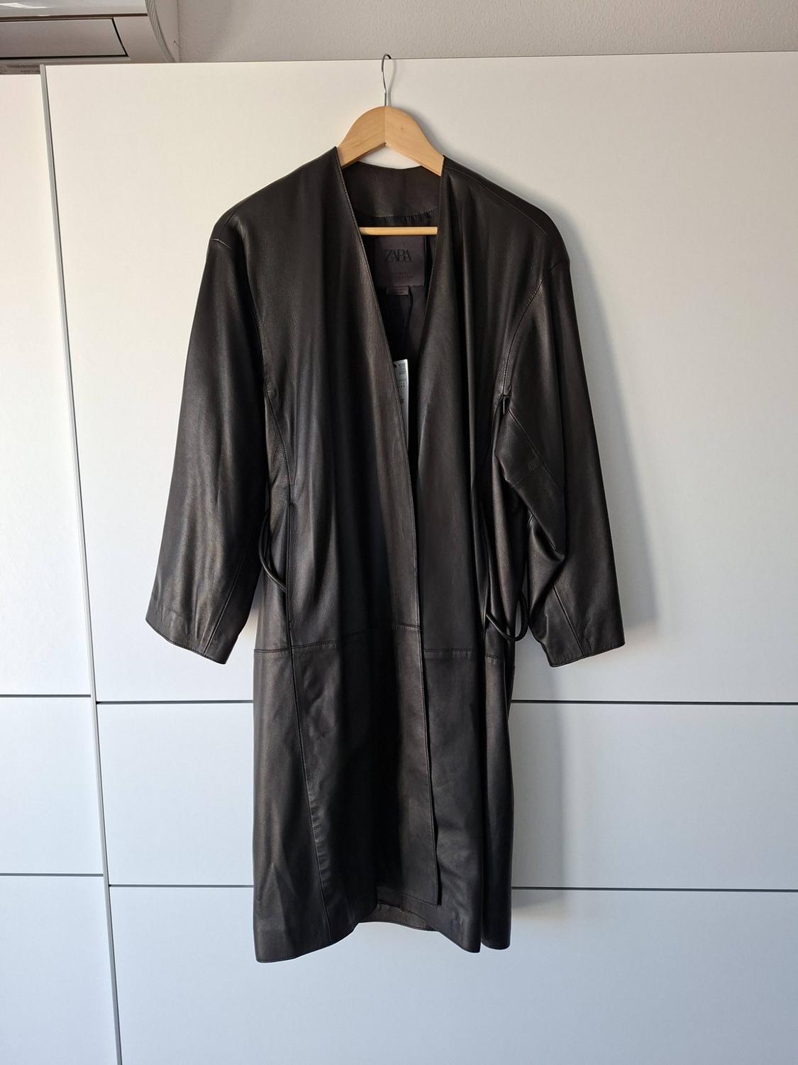 Long black collarless leather coat hanging on a wooden hanger in front of a white wardrobe. Label with the text ZARA.