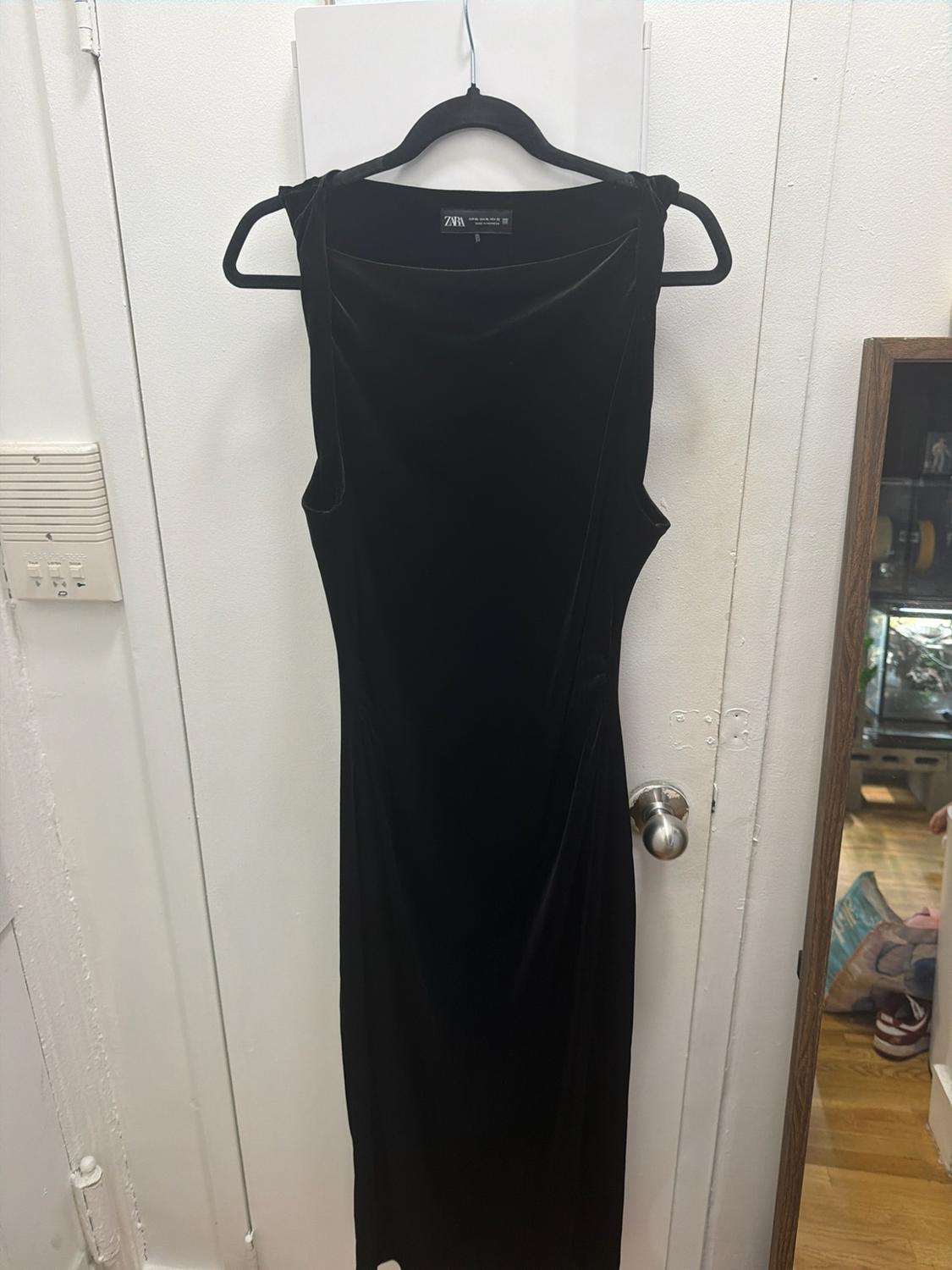 Sleeveless black dress hanging from a hanger on a white door. Tag with the text ZARA.