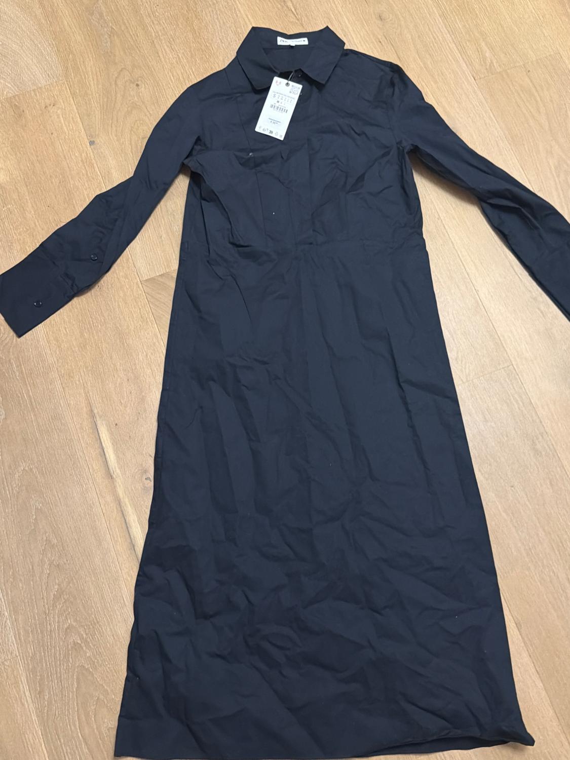 Long-sleeved black dress with a classic collar on a wooden floor. Visible tag with small text hanging from the collar.