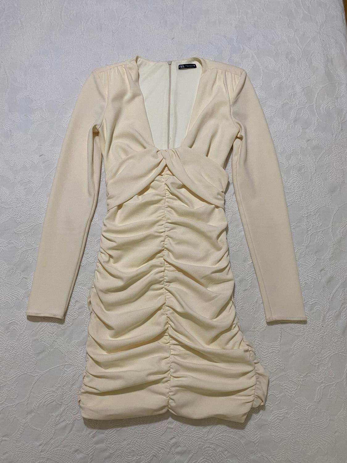 Beige long-sleeved dress with ruching and a V-neckline, on a white surface with a floral texture.