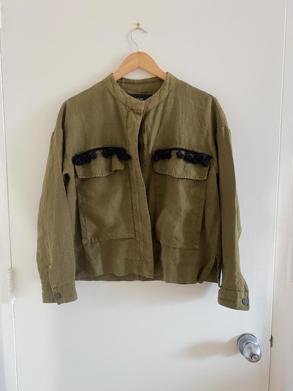 Olive green jacket hanging on a wooden hanger, with black fringe details on the front pockets, over a white door.