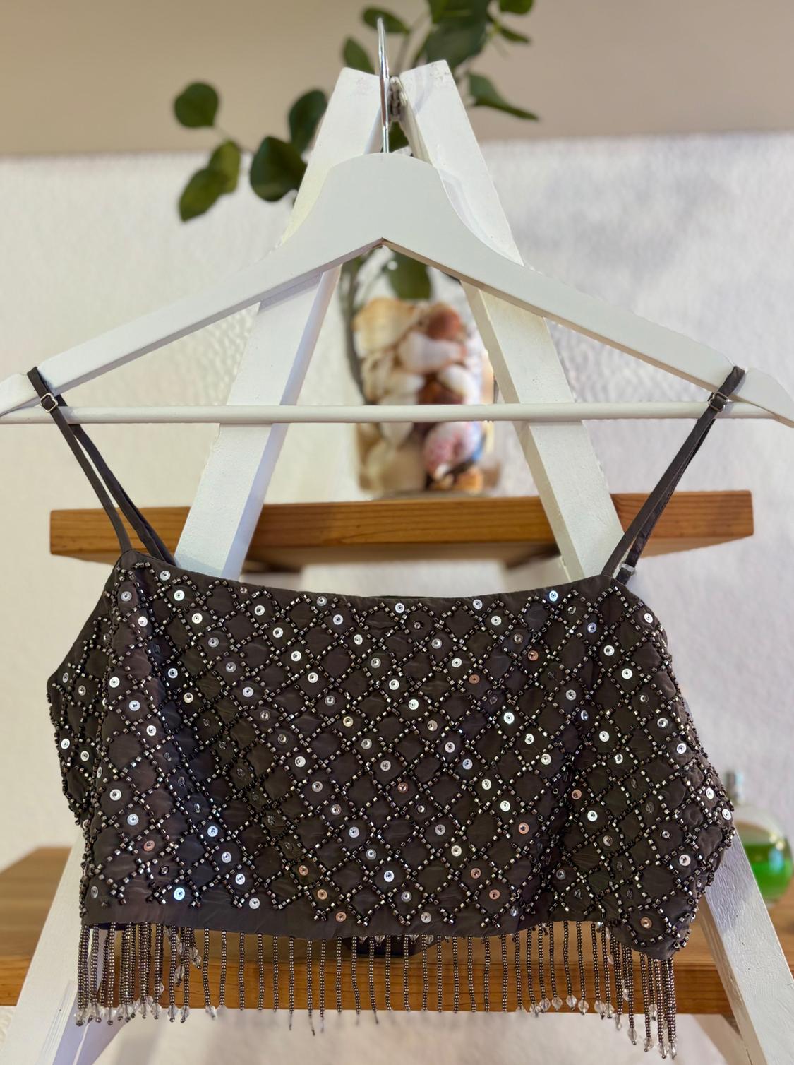 Black crop top with thin straps, embellished with sequins and fringe, hanging on a white hanger. Background with plants and shells.