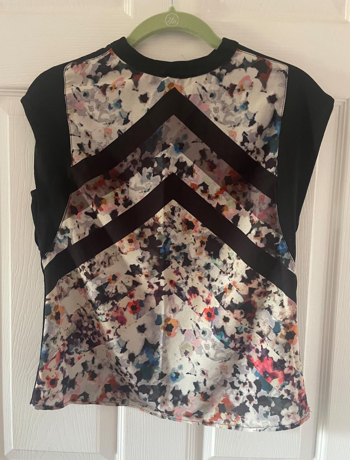 Sleeveless blouse with an abstract floral print and black zigzag stripes, hanging on a green hanger in front of a white door.