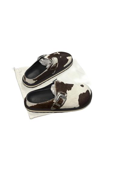 A pair of cow-print clogs with silver buckles, sitting on a white fabric bag. The name of the brand ZARA can be seen on the insole.