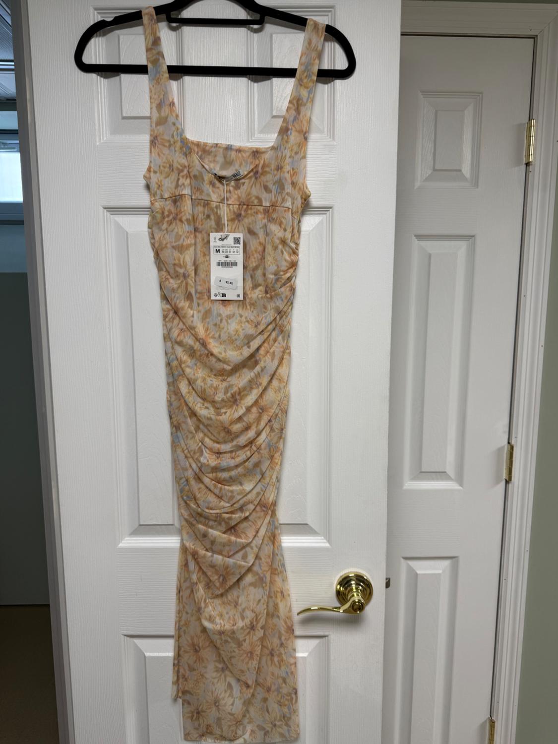 Long floral print dress in pastel shades, hanging on a white door. Visible label with size M and price.