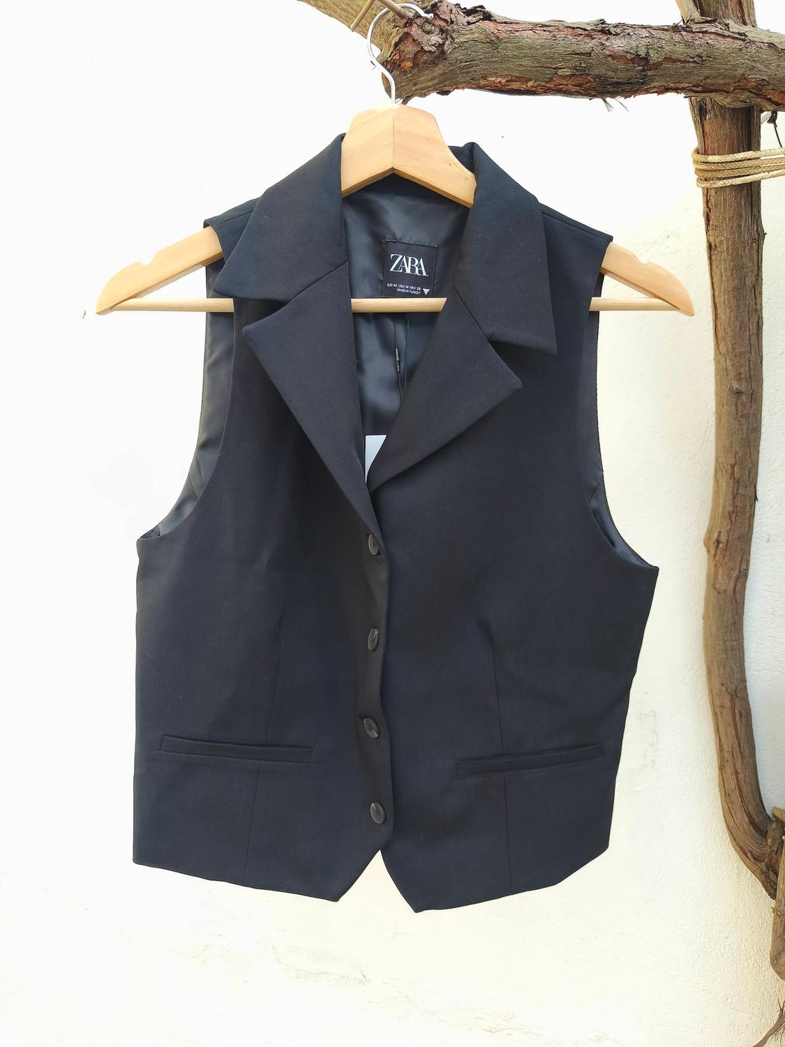Black sleeveless waistcoat hanging on a wooden hanger, with a white background and tree branch. Visible label: "ZARA".