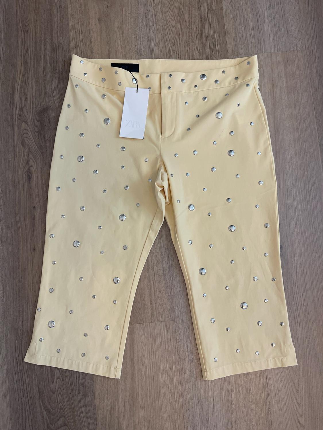 Yellow capri pants decorated with silver studs, with a ZARA brand label, presented on a wooden floor.