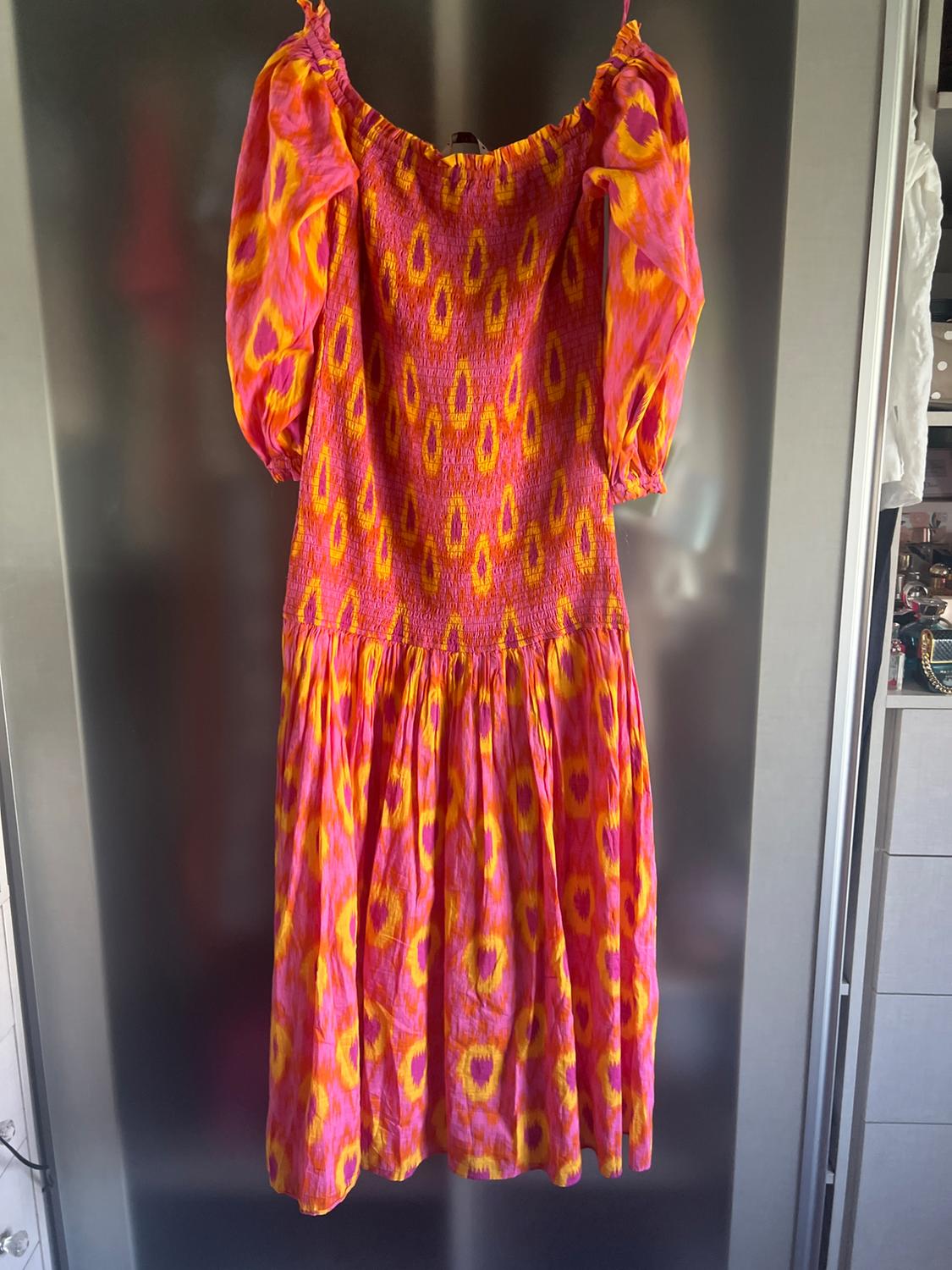 Long pink and orange patterned dress with a shirred bodice and puff sleeves, hanging in front of a metallic surface.